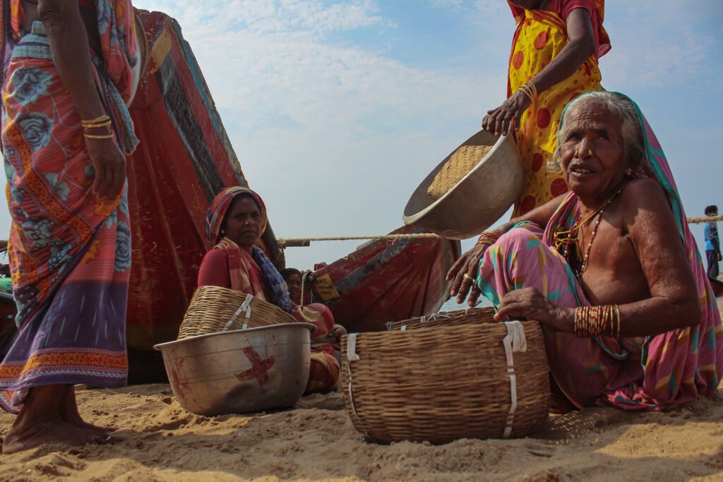 A group of women dressed in colorful saris sit together on the sand. They are gathered around woven baskets, sorting through the day's catch.