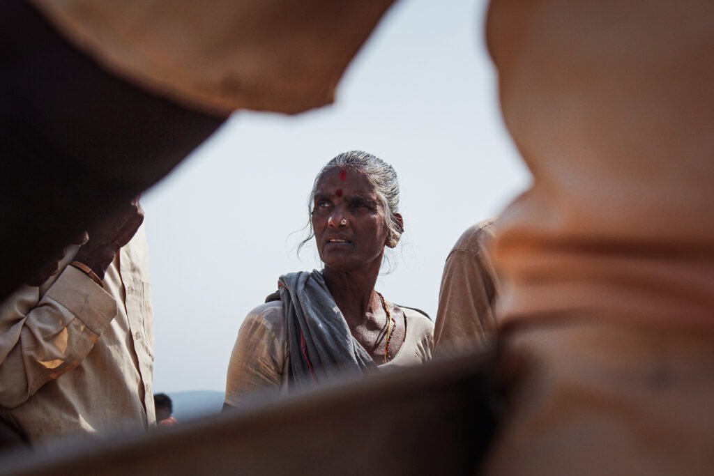 A close-up portrait of a woman looking off-camera, with a red bindi on her forehead. Her face is framed by the blurred, out-of-focus arms and clothing of people in the foreground.