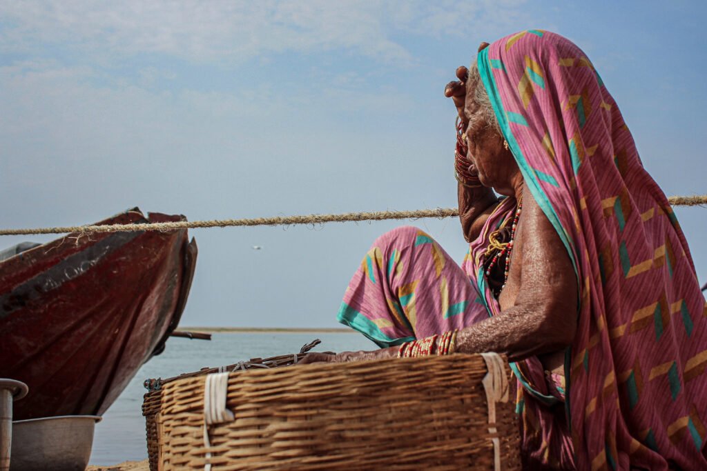An older woman wearing a vibrant pink and green sari sits on the sand beside a wooden boat. She rests her hand on her head, looking thoughtfully out toward the ocean.