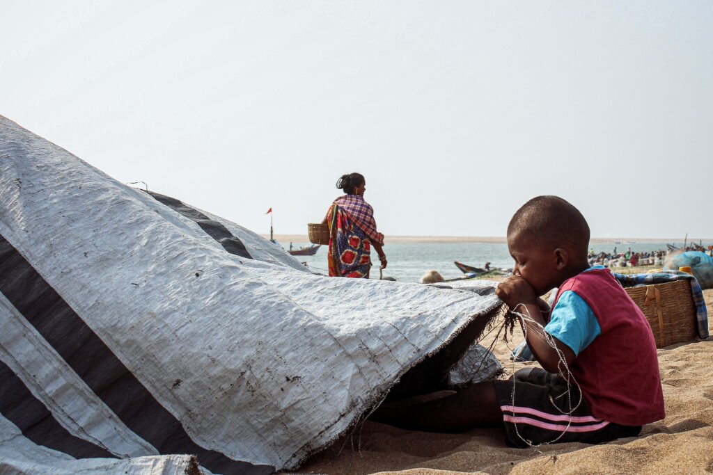 A young child sits quietly on the sand, partially shaded by a large, draped tarp. Boats and fishermen are visible in the bright, distant background of the beach.