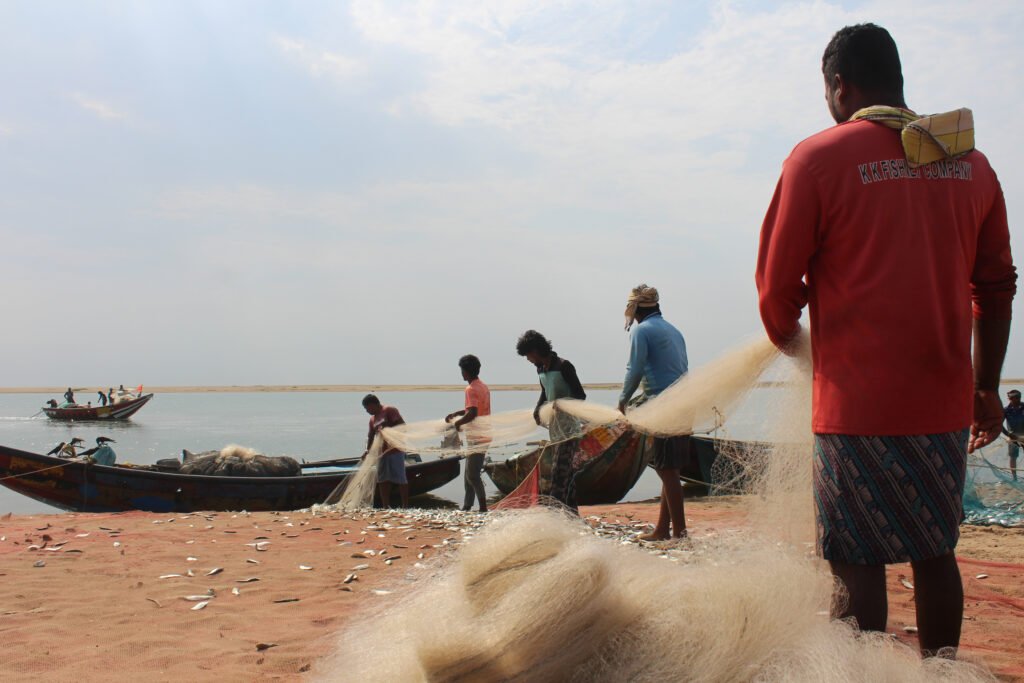 A fisherman in a bright red shirt stands on a sandy beach, pulling a large white fishing net. Other fishermen and boats are visible in the background against a clear sky.