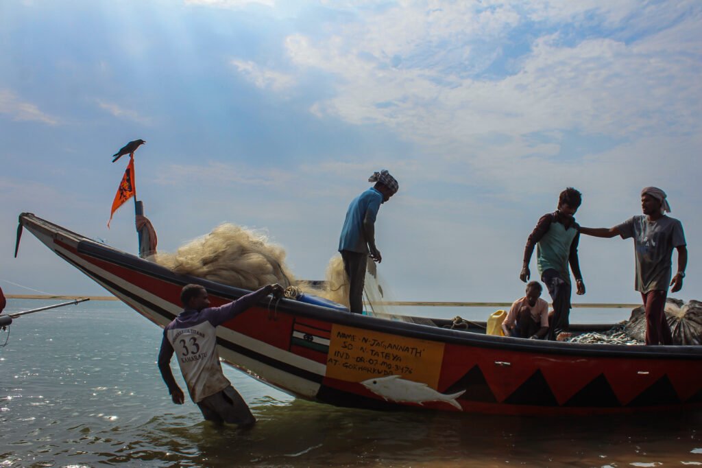 Several fishermen balance on a painted wooden boat actively bobbing in the water. They are hauling nets over the side, with a small saffron flag flying from the bow.