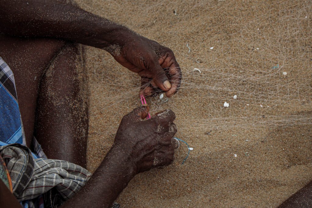 A tight, macro shot of a fisherman's hands resting on the granular sand. The hands are weathered and actively tying or repairing a fine, translucent fishing net.