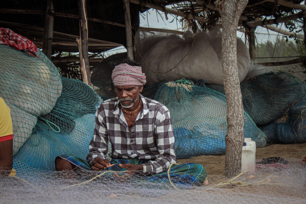A man in a plaid shirt sits on the sand under a makeshift shelter made of wooden poles and fabric. He is surrounded by fishing nets and looks directly and intently into the camera.