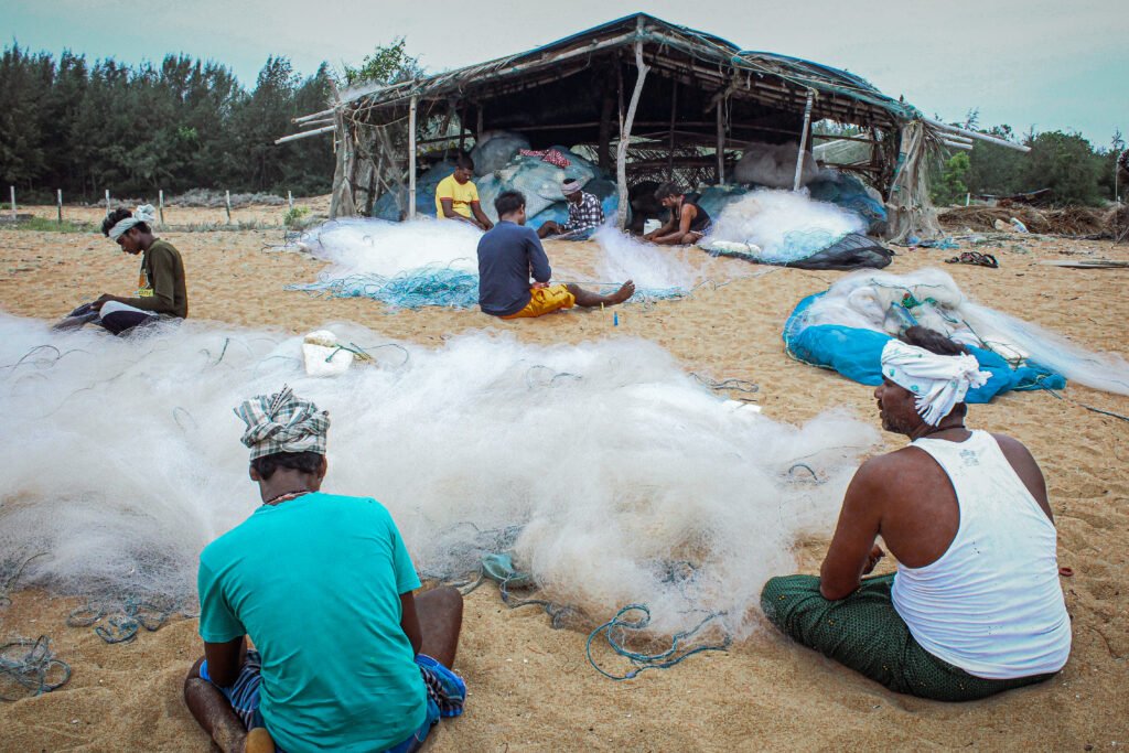 A wide shot of a bustling beach scene with fishermen working under a large, shaded shelter. Piles of white fishing nets are spread out across the sand as the men organize their gear.